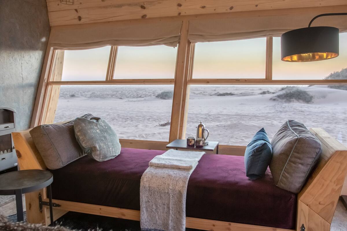 A cozy room with a wooden bed, a small table with a kettle, and a view of the beach through large windows.