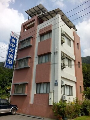 Un edificio de varios pisos con un letrero que indica que es un hotel. Cuenta con múltiples ventanas en cada planta y un techo plano con un panel solar.