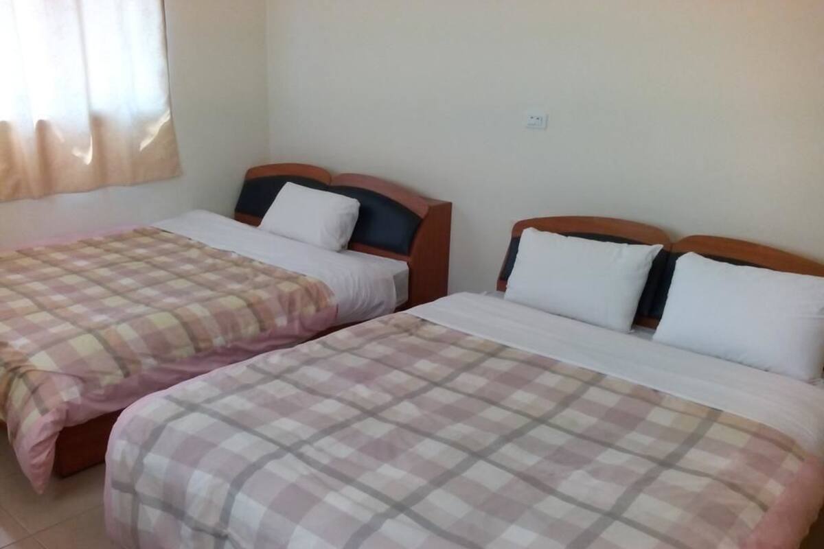 Two single beds with wooden headboards, a window with curtains, and a plain wall with a power outlet.