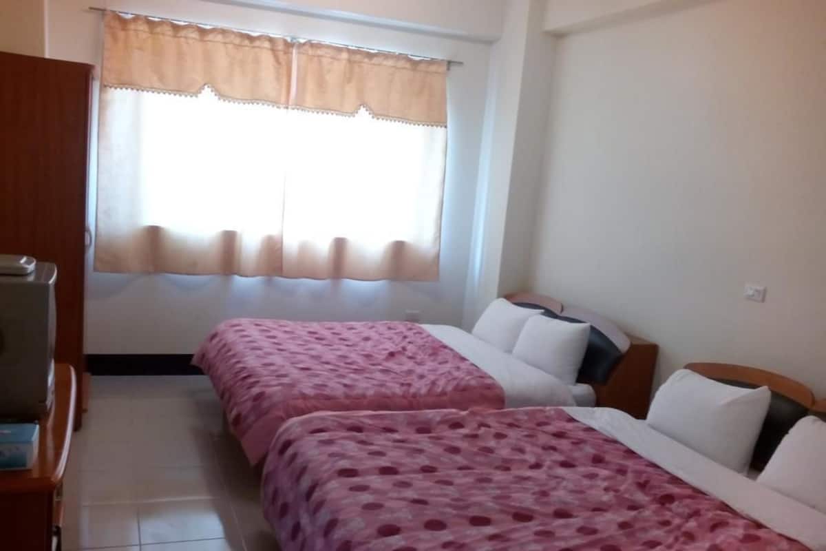 A room with two beds, a wooden wardrobe, a window with a beige curtain, and a tiled floor.