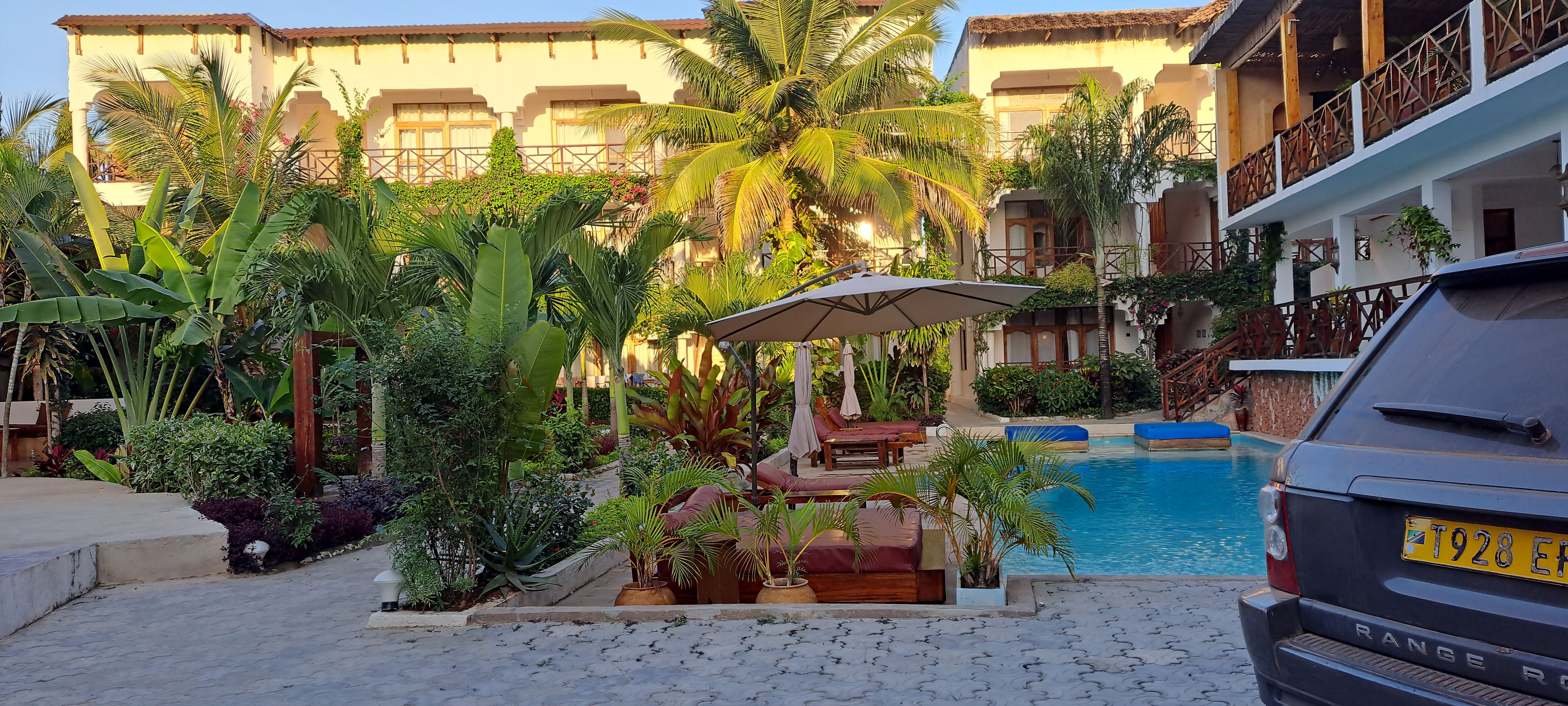 A pool area with lounge chairs and umbrellas, surrounded by lush tropical plants and a building with balconies.