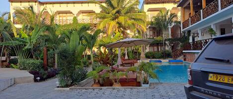 A pool area with lounge chairs and umbrellas, surrounded by lush tropical plants and a building with balconies.