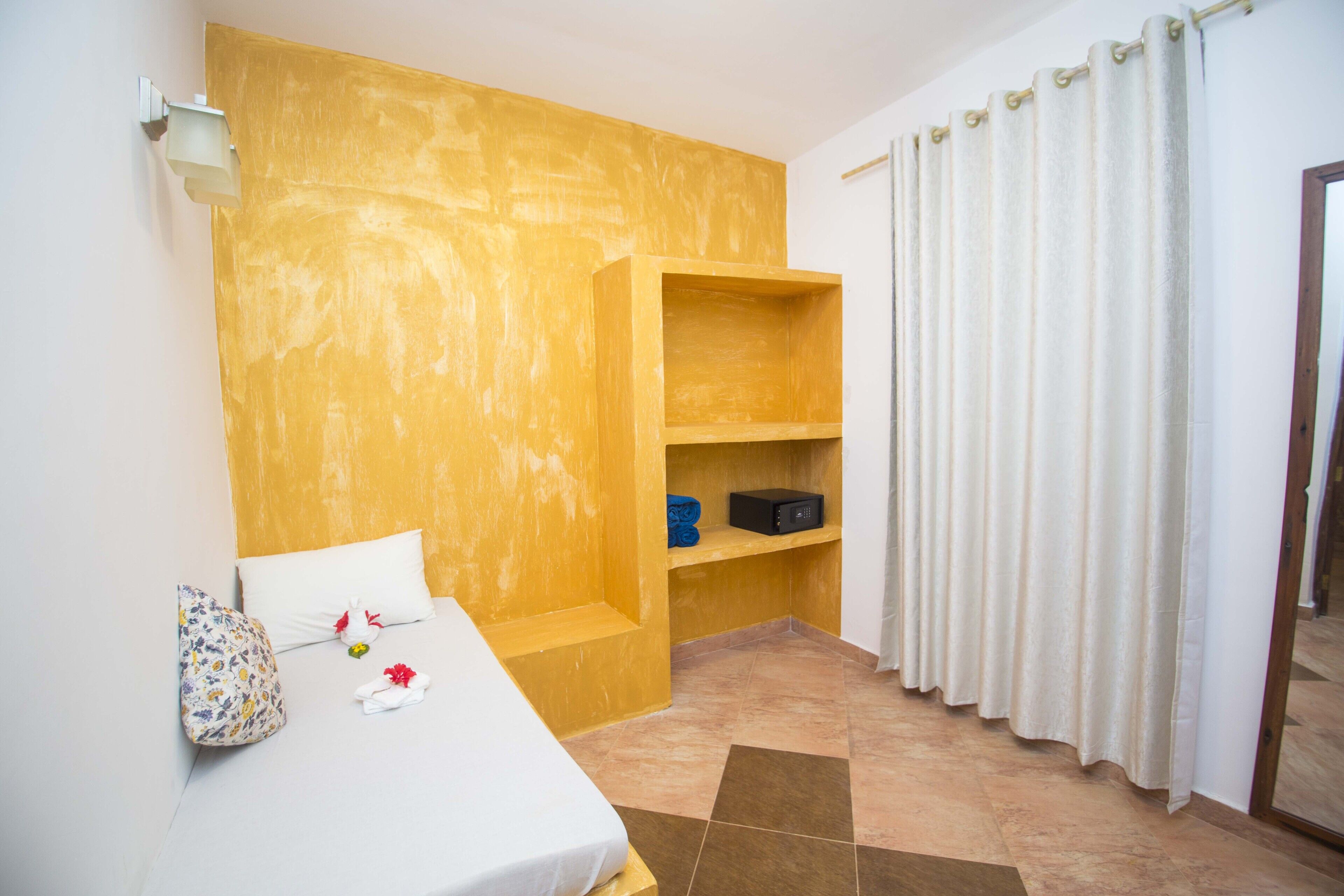Une chambre d’hôtel avec un lit, une étagère en bois, un rideau blanc et un sol carrelé.