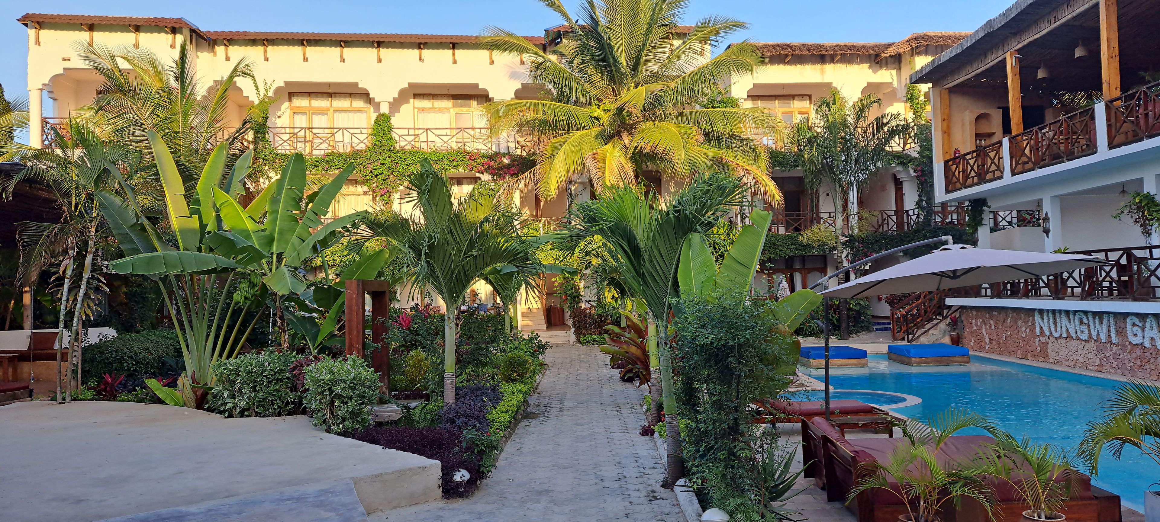 Pátio de hotel com piscina, cercado por plantas tropicais exuberantes e um edifício com varandas.