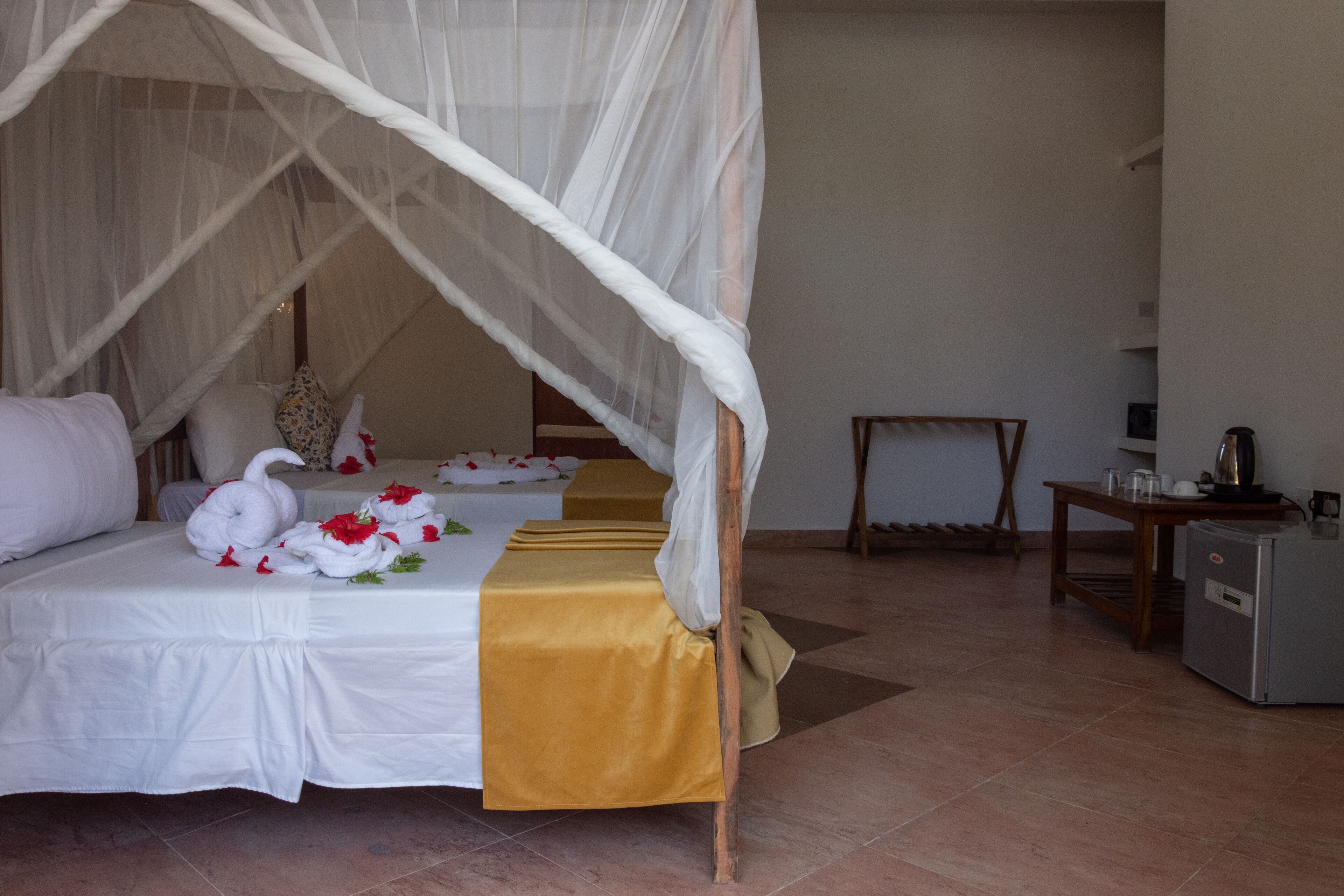 Une chambre d’hôtel avec un lit recouvert de draps blancs, un baldaquin et des cygnes décoratifs en tissu. On y trouve une petite table avec une bouilloire et des tasses, ainsi qu’un réfrigérateur.