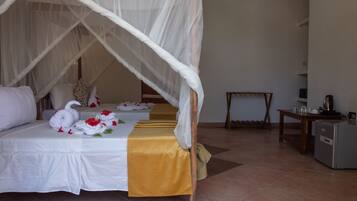 Une chambre d’hôtel avec un lit recouvert de draps blancs, un baldaquin et des cygnes décoratifs en tissu. On y trouve une petite table avec une bouilloire et des tasses, ainsi qu’un réfrigérateur.
