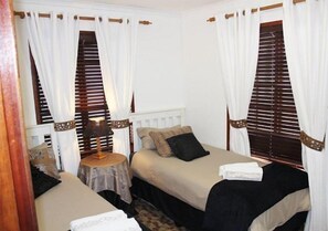 Een kamer met twee bedden, elk met een houten hoofdbord, witte gordijnen en een klein rond tafeltje met een lamp.