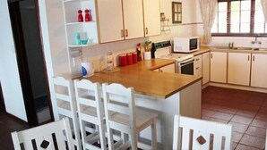A compact kitchen with white cabinets, a wooden countertop, and a dining area with white chairs. - 303 Kusweg (Great Brak River)