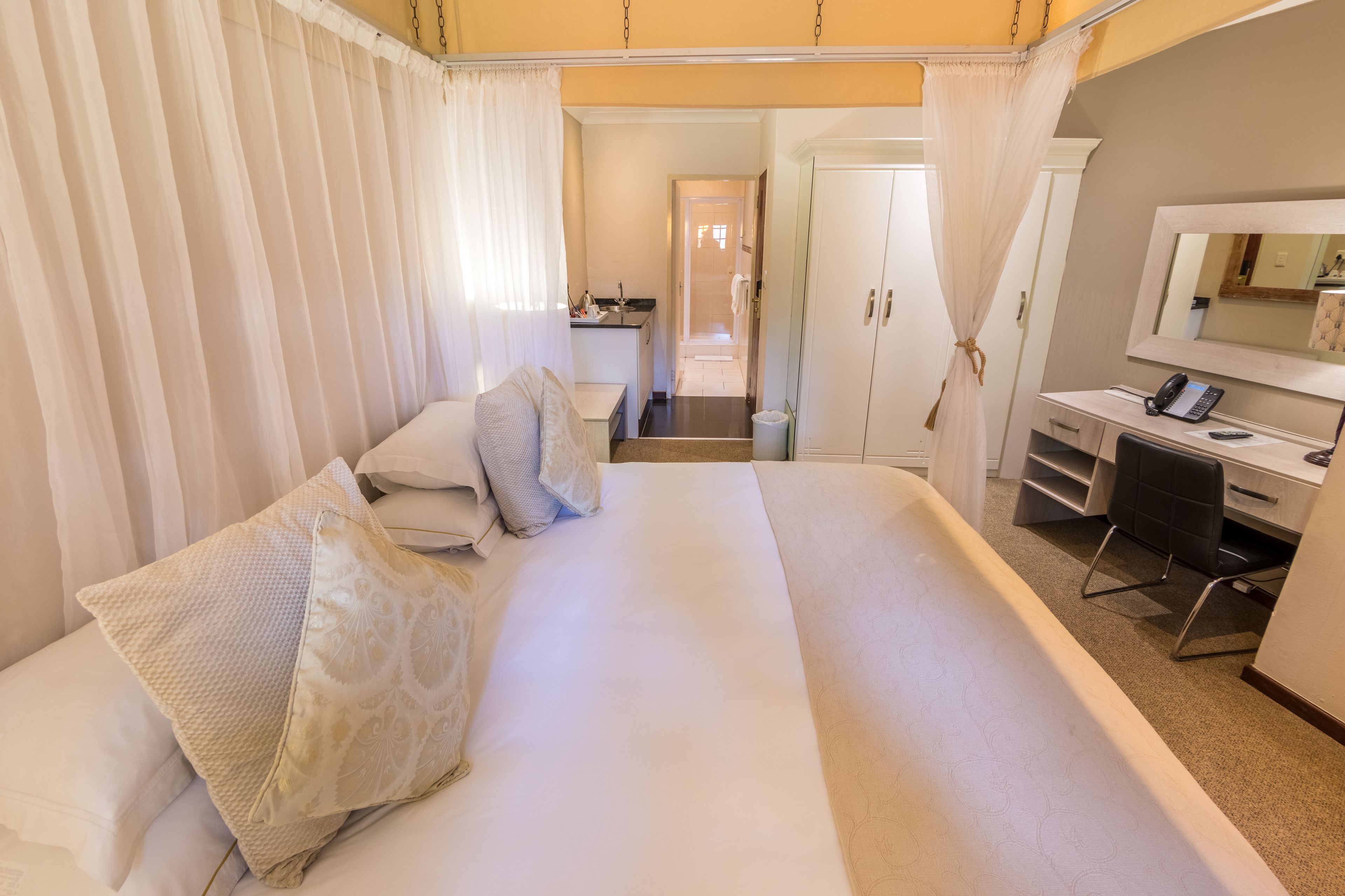 Une chambre d’hôtel avec un grand lit, un bureau avec une chaise, un miroir et, au fond, une petite kitchenette.