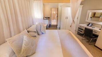 Une chambre d’hôtel avec un grand lit, un bureau avec une chaise, un miroir et, au fond, une petite kitchenette.