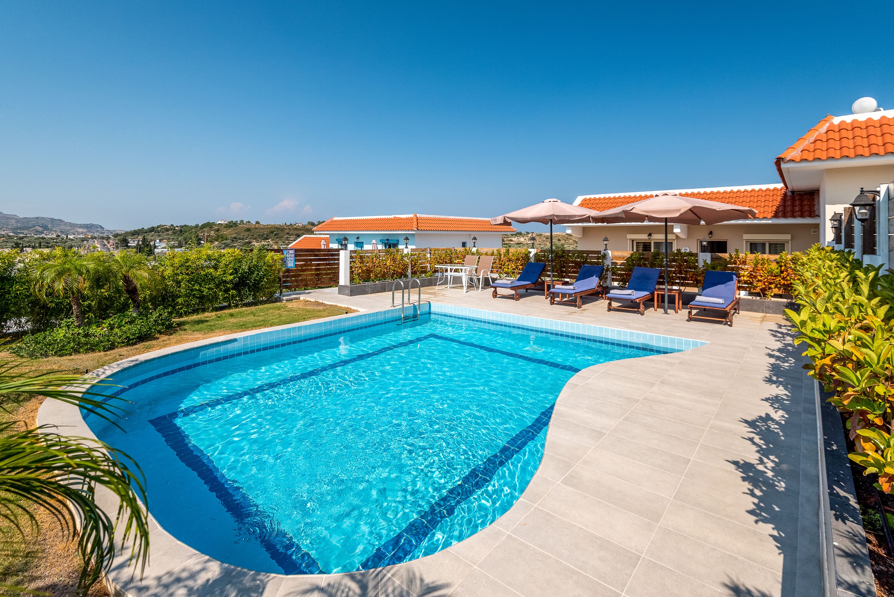 Una piscina con sillones de playa y sombrillas, rodeada de un jardín ajardinado y edificios con tejados de tejas rojas.