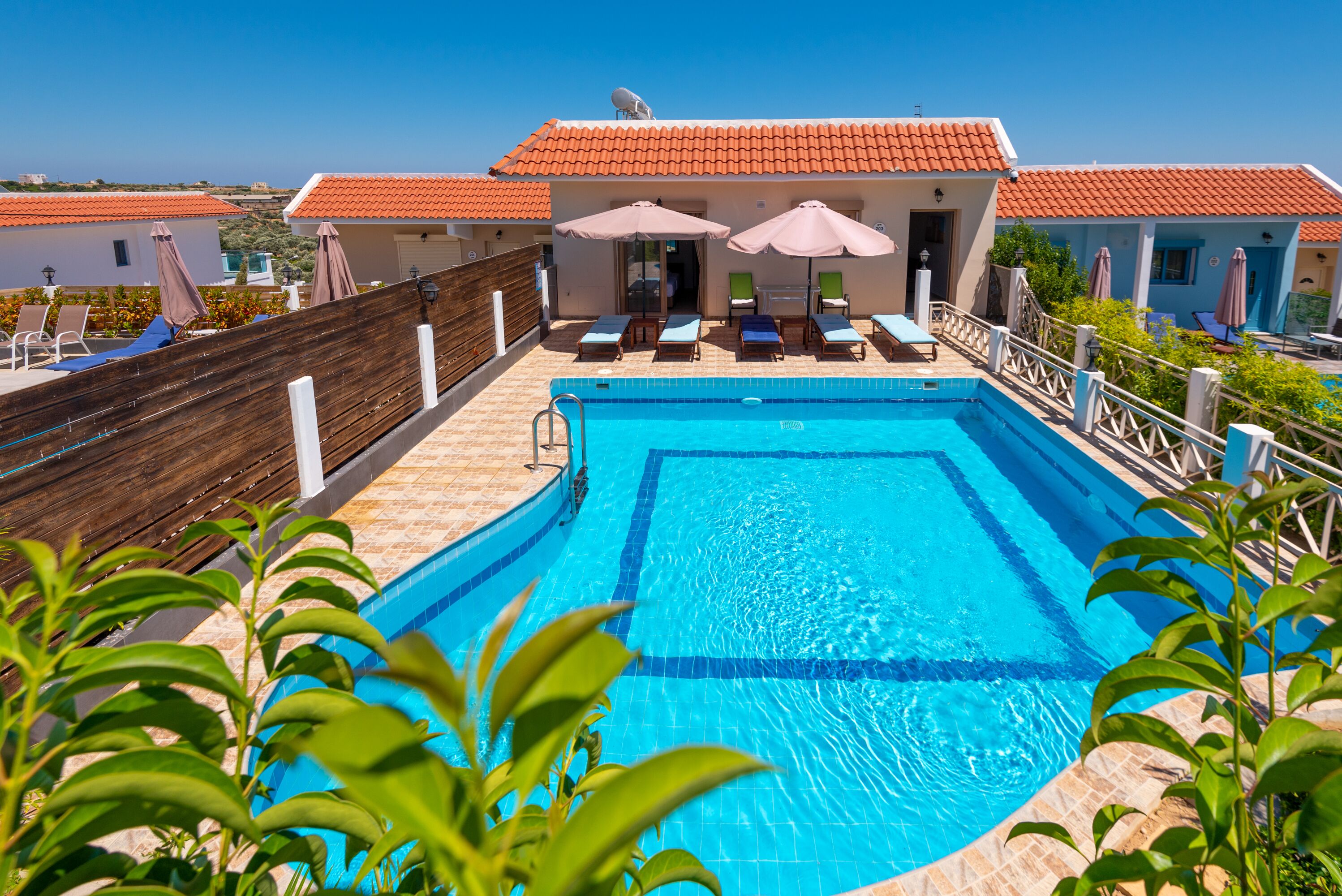 A swimming pool with lounge chairs and umbrellas, surrounded by a wooden fence and a building with a red-tiled roof.