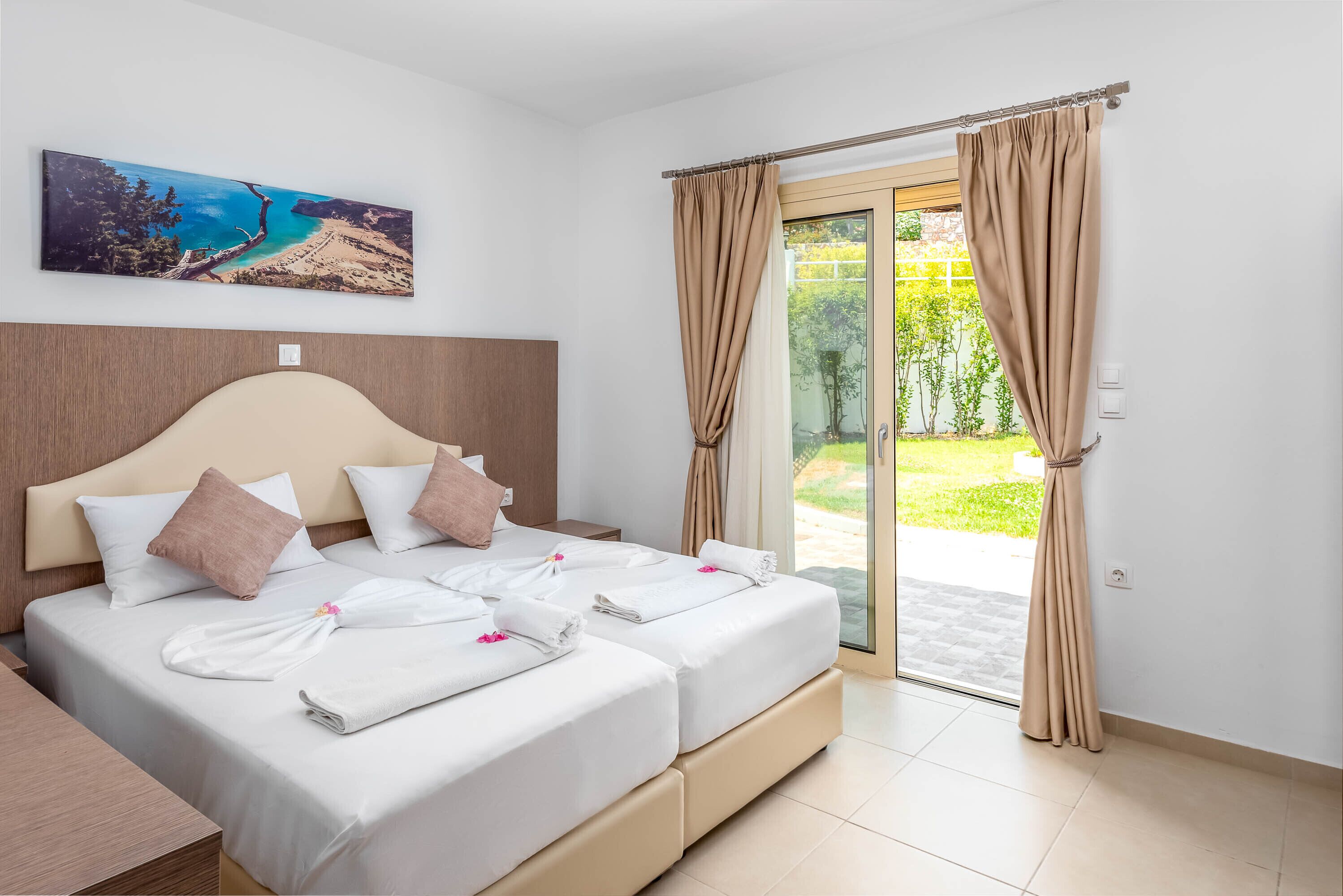 Une chambre d’hôtel avec deux lits, une grande fenêtre avec des rideaux, un tableau représentant une plage et une vue sur la verdure à l’extérieur.
