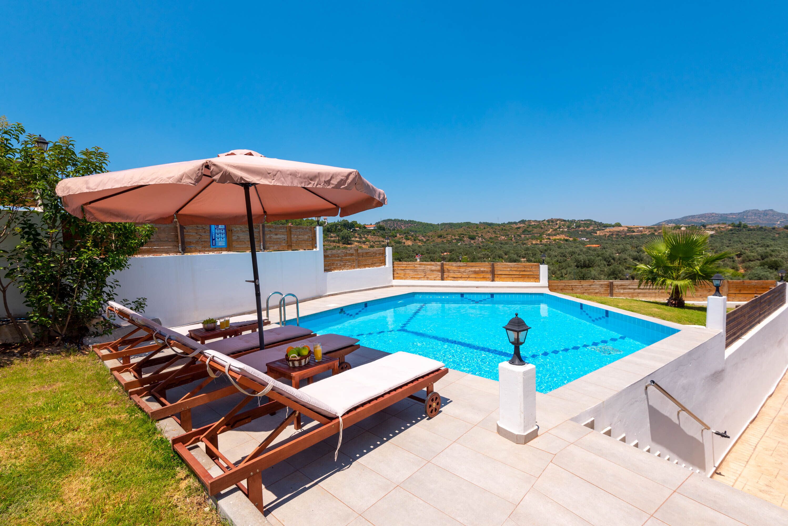 A swimming pool with lounge chairs and an umbrella, surrounded by a white wall and a view of hills.