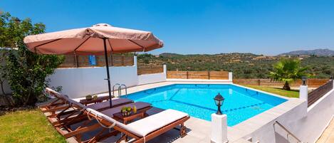 A swimming pool with lounge chairs and an umbrella, surrounded by a white wall and a view of hills.
