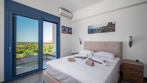 Un dormitorio con una cama grande, una mesita de noche, un mueble cajonero de madera y vistas al verdor a través de un ventanal corredero.