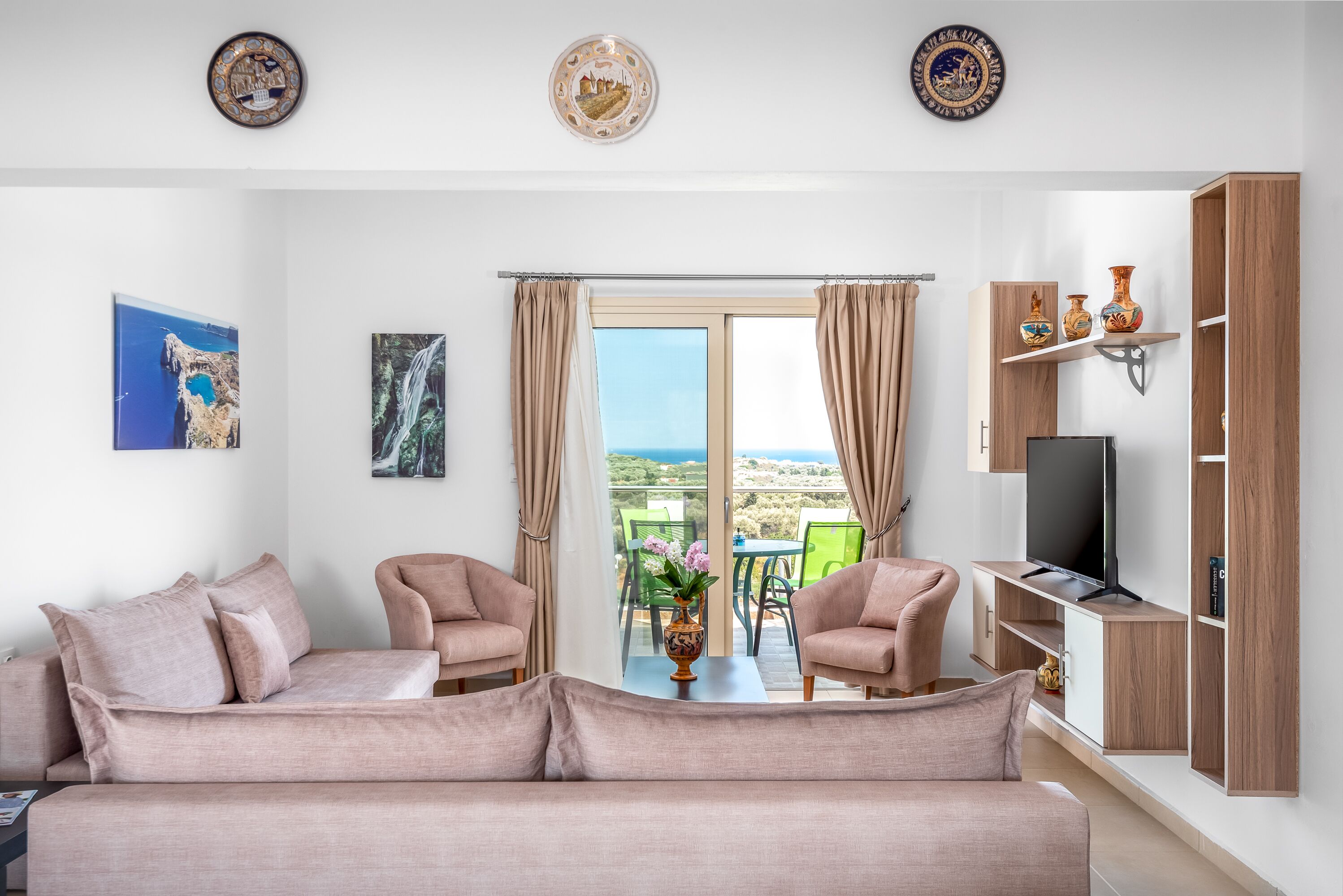 Una sala de estar con sofá, dos sillones, una mesa de centro y un mueble para televisión. Hay dos ventanas con cortinas, vista al exterior y una pared decorada con dos placas.