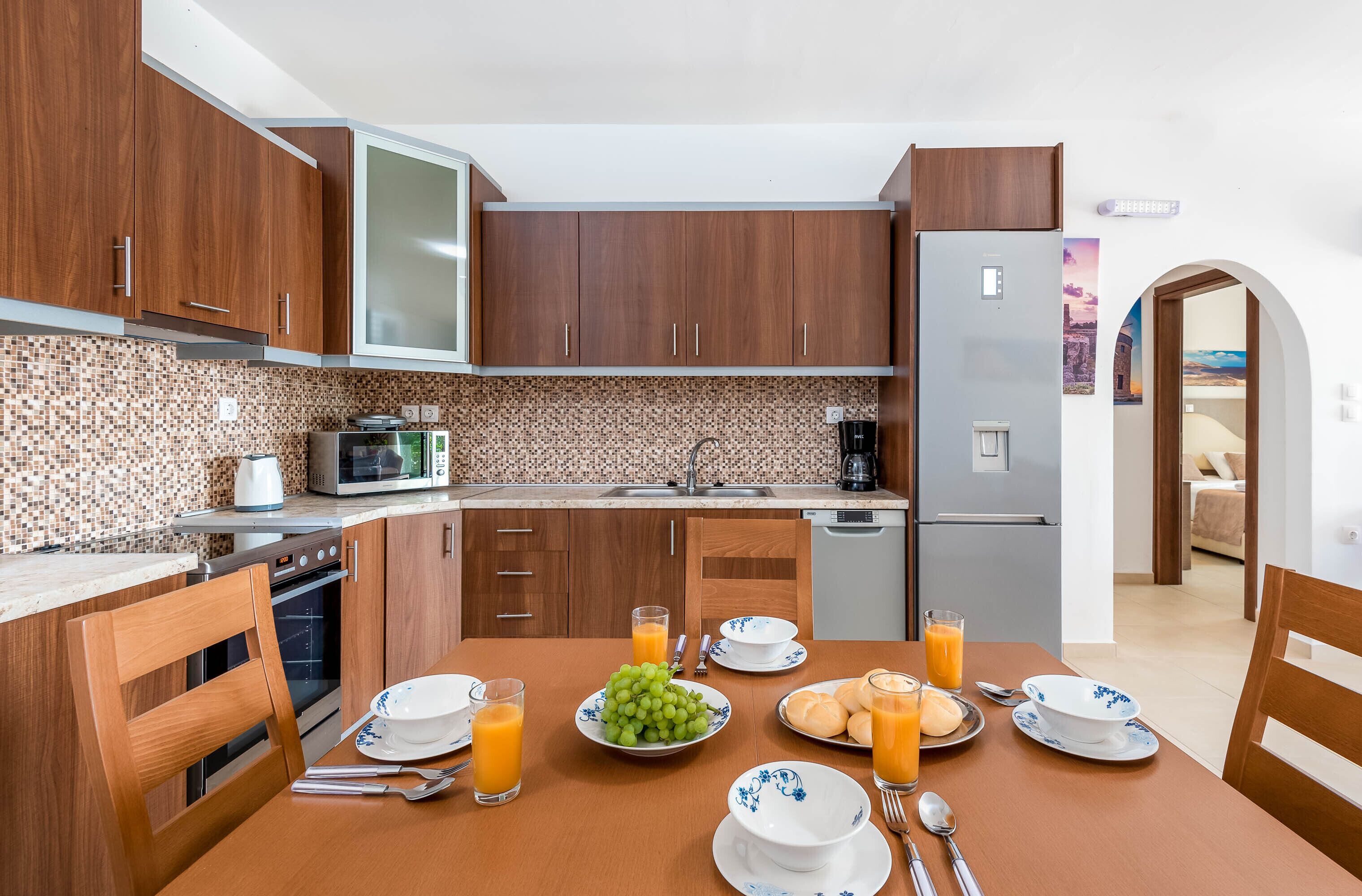 Una cocina moderna con armarios de madera, microondas y refrigerador. Hay una mesa de comedor preparada con platos, tazones y vasos de jugo de naranja.