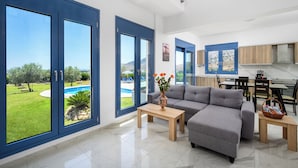Una sala de estar moderna con un sofá gris, una mesa de centro de madera y vistas a una piscina y un jardín.