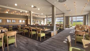 A modern restaurant with wooden tables and chairs, large windows offering a view of the outdoors, and a variety of hanging lights.