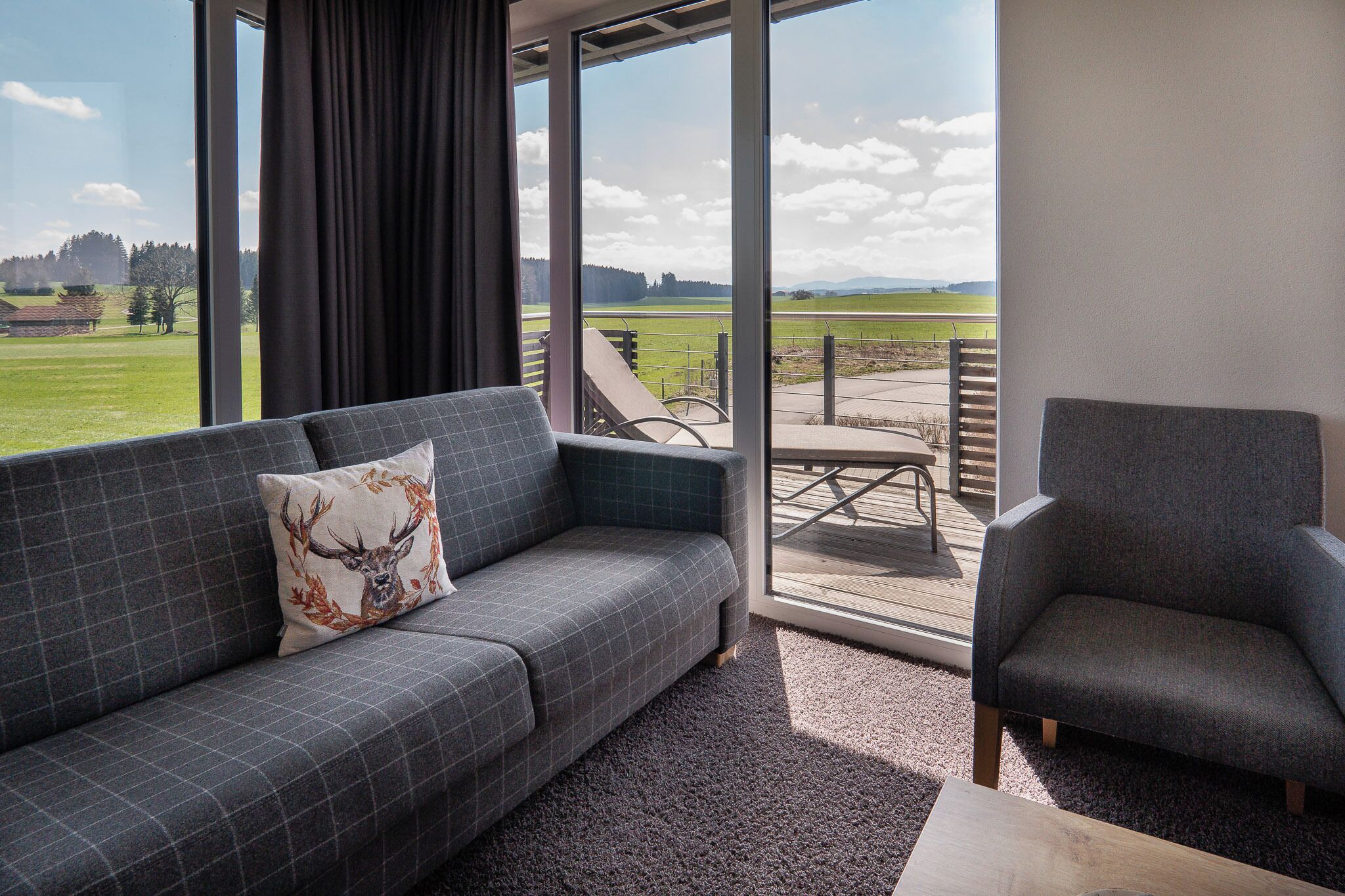 A modern living room with a grey sofa, a matching armchair, and a large window offering a view of a green field and distant hills.