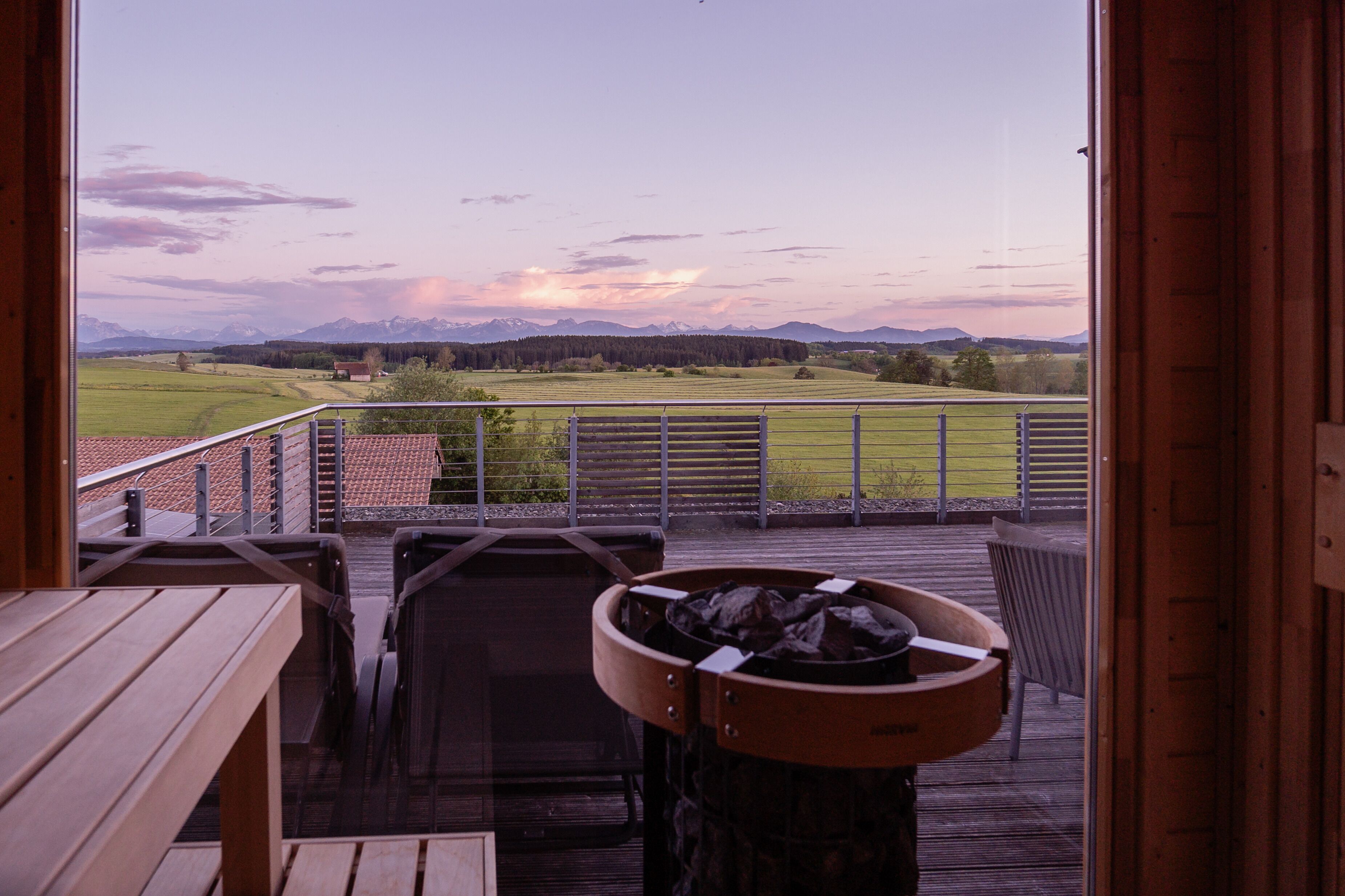 Une terrasse en bois avec un banc et un foyer, donnant sur un champ verdoyant et des montagnes lointaines.