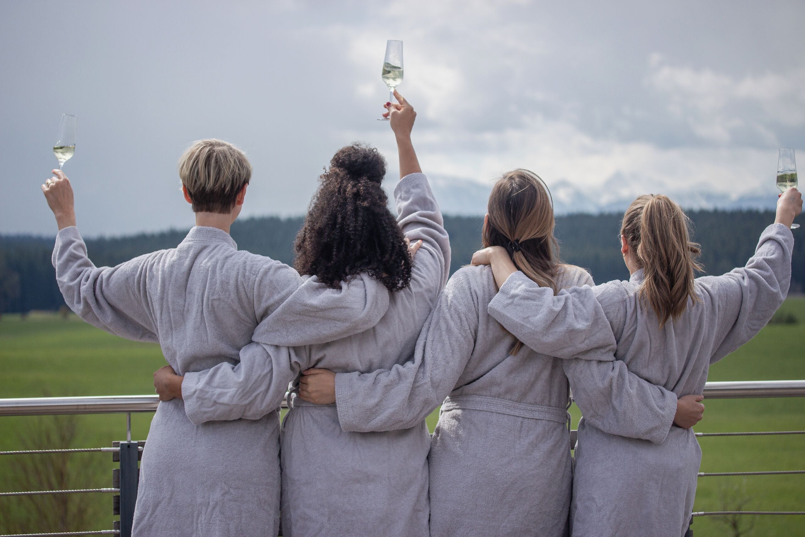 Four people in grey robes standing on a balcony with a view of a green landscape and mountains in the distance.