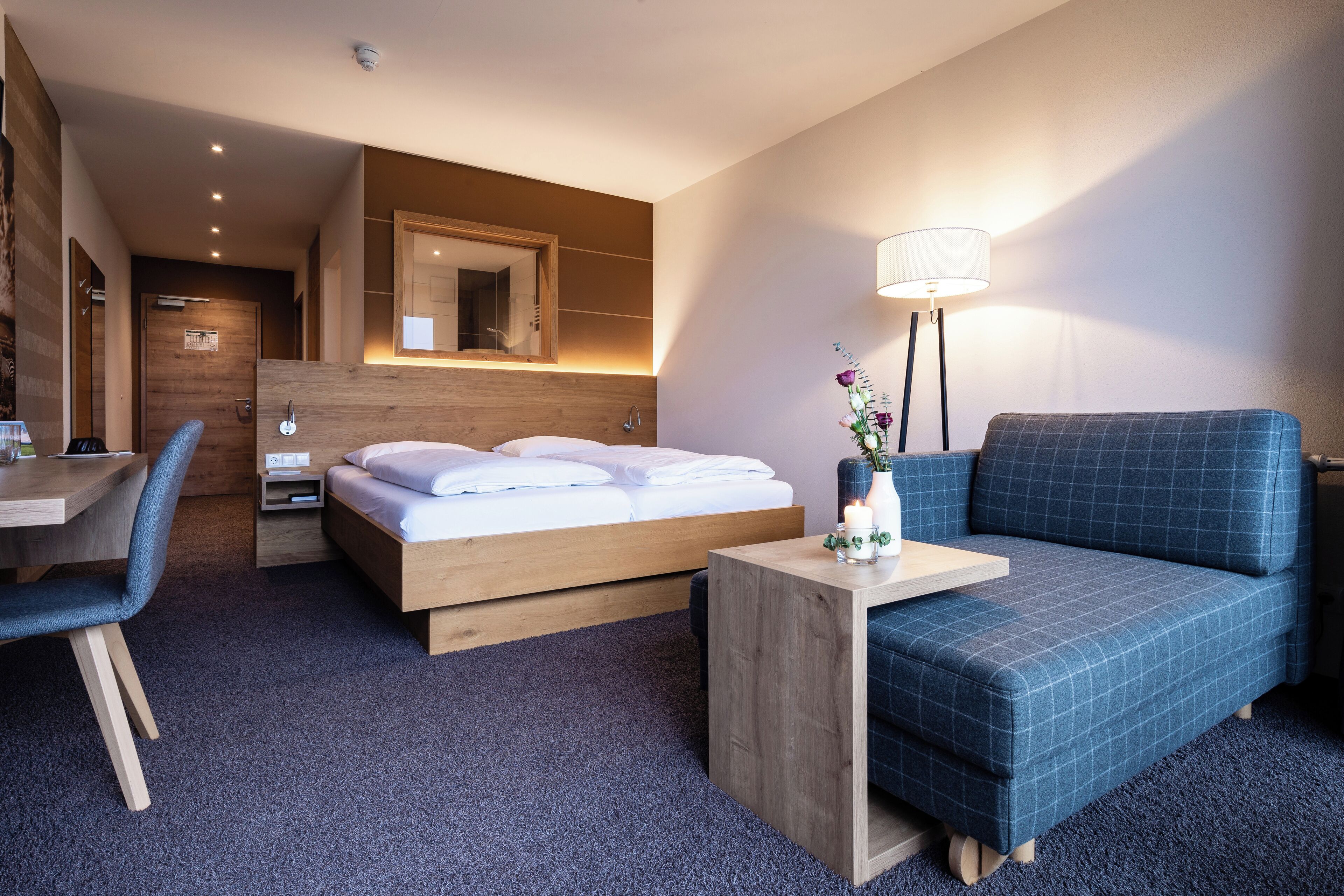 Une chambre d’hôtel moderne équipée d’un lit, d’un bureau, d’une chaise, d’un canapé et d’une table de chevet avec une lampe.