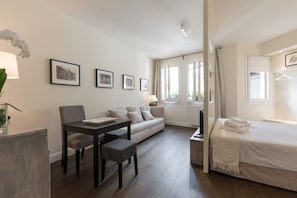 A modern hotel room with a bed, a sofa, a dining table, and a desk. There are framed pictures on the wall, a window with curtains, and a plant on the desk. - Suite & View (Florence)