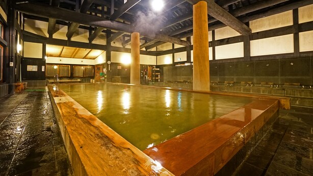 A traditional Japanese onsen with a wooden bath, stone walls, and a high ceiling with exposed beams.