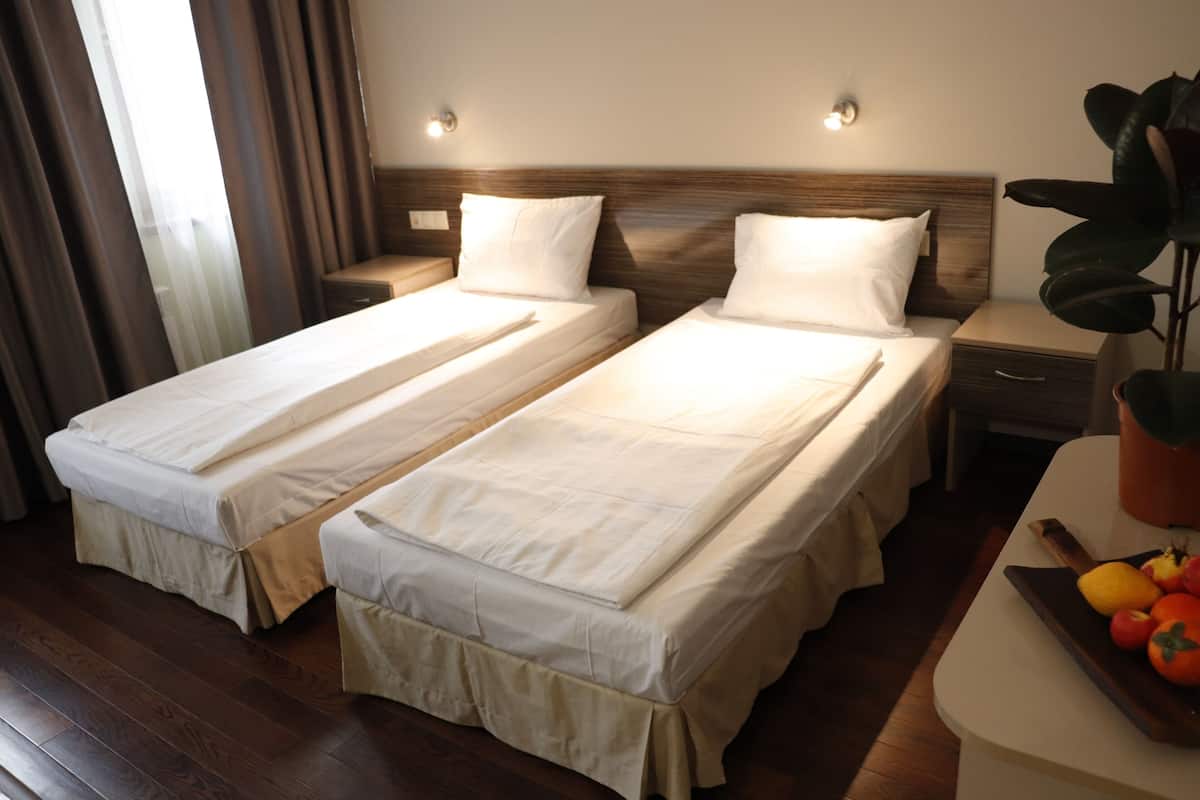 Quarto de hotel com duas camas de solteiro, piso de madeira, mesinhas de cabeceira, uma plantadeira e um tabuleiro de frutas.