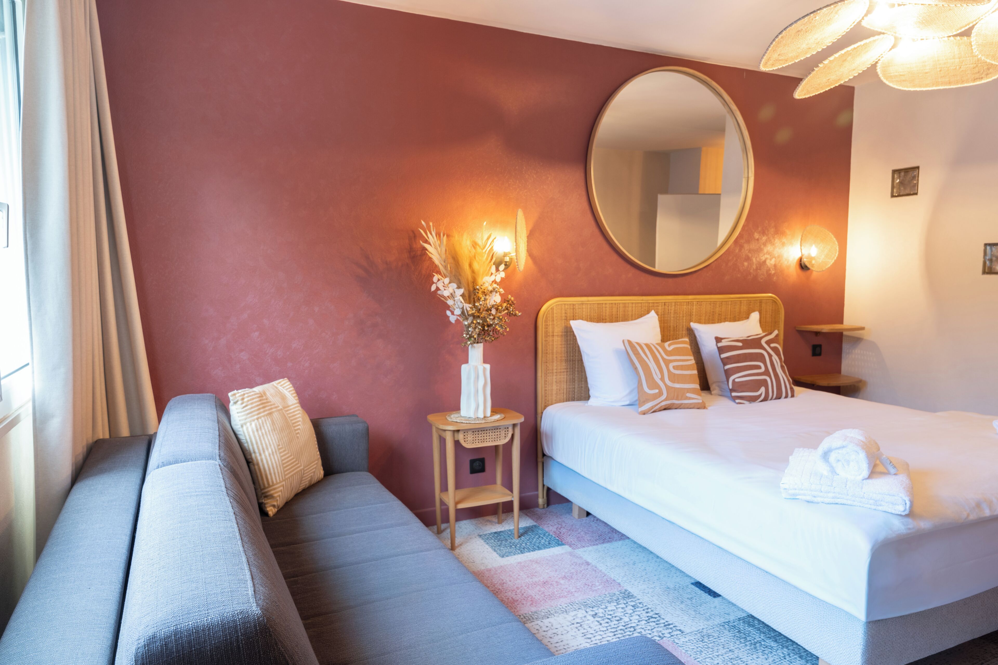Habitación de hotel con cama, sofá, espejo redondo, una pequeña mesa con un florero y una lámpara de pared.