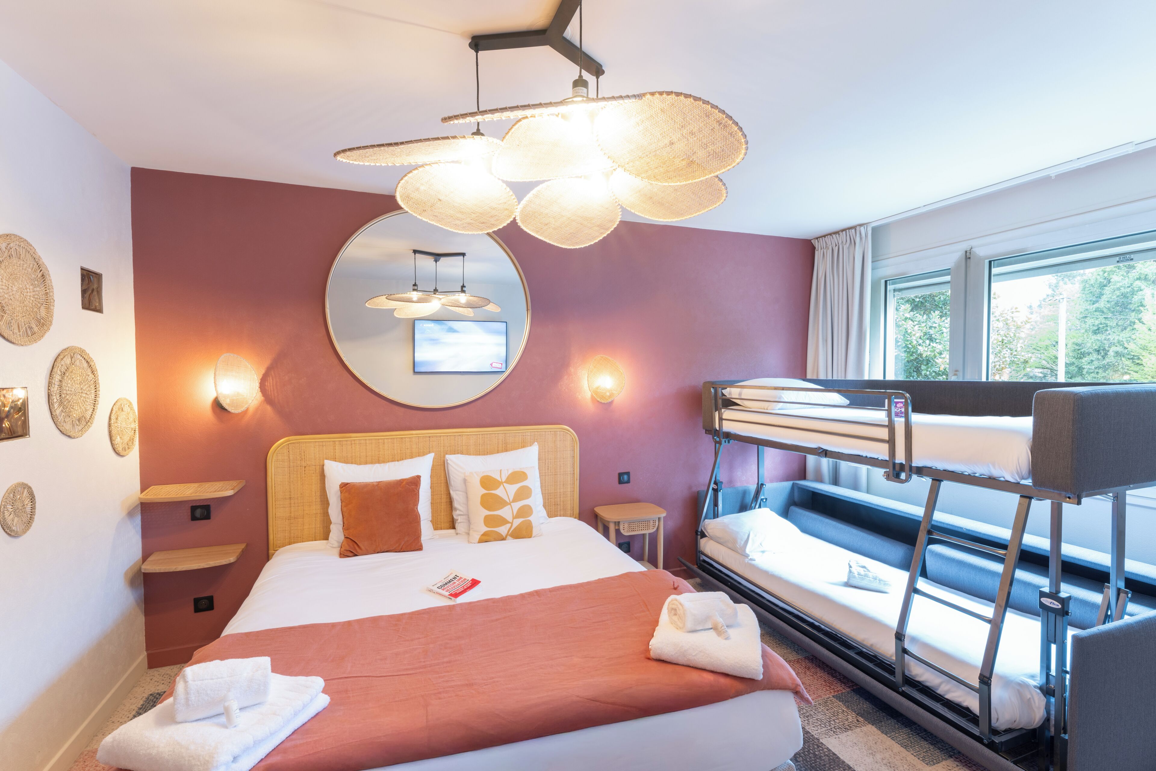 Ein Hotelzimmer mit Etagenbett, einem großen Bett mit roter Decke, einem runden Spiegel, Wandleuchten und einem Fenster mit Vorhängen.