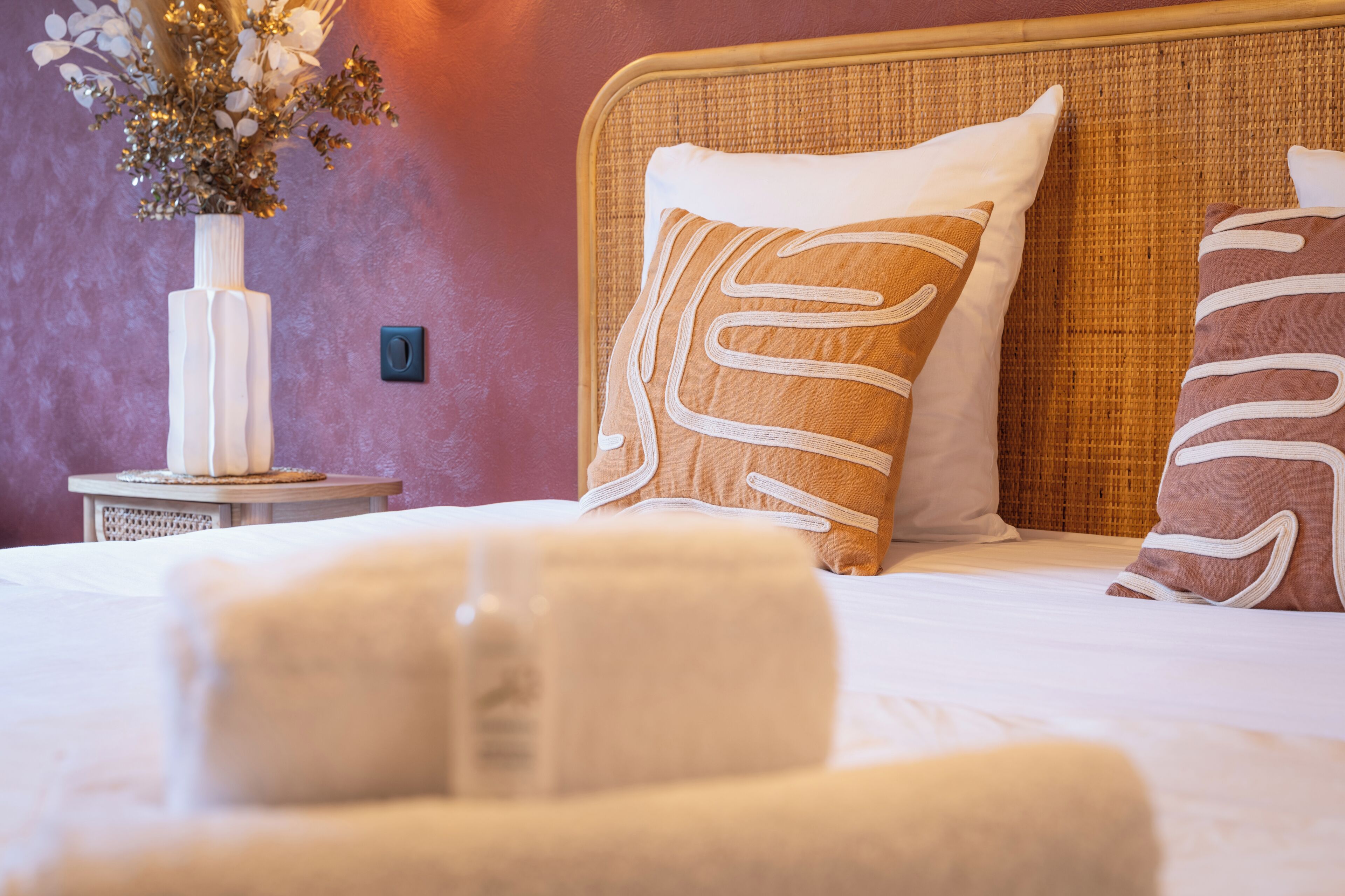 A bed with white linens, two pillows with white and brown patterns, a vase with flowers, and a small table with a candle.