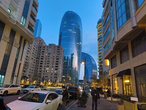 A cityscape with modern buildings, a curved glass skyscraper, and a busy street with cars and pedestrians. - Deluxe City Hotel (Baku)