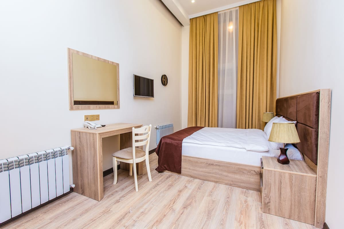 A hotel room with a wooden desk, chair, and bed. There is a TV mounted on the wall, a bedside table with a lamp, and a window with curtains.
