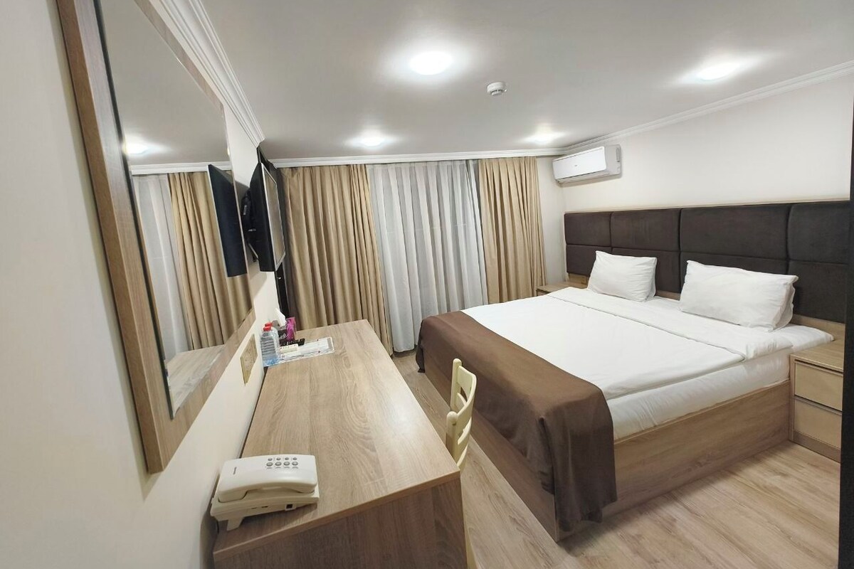 A hotel room with a bed, bedside table, chair, desk, mirror, and a wall-mounted air conditioning unit.