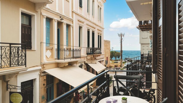 A balcony with a table and chairs, overlooking a street with buildings and a view of the sea. - Elia Palazzo Hotel (Chania)