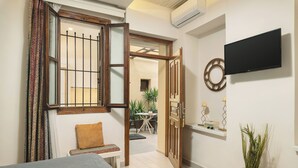 A hotel room with a bed, a bench, a small table, a potted plant, and a TV mounted on the wall. - Elia Palazzo Hotel (Chania)