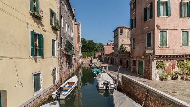 Un canal estrecho con barcos amarrados a ambos lados, bordeado por edificios con contraventanas verdes y balcones.