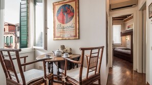 Un comedor con sillas de madera y una mesa, un cartel enmarcado en la pared, y vista a un dormitorio por una puerta abierta.