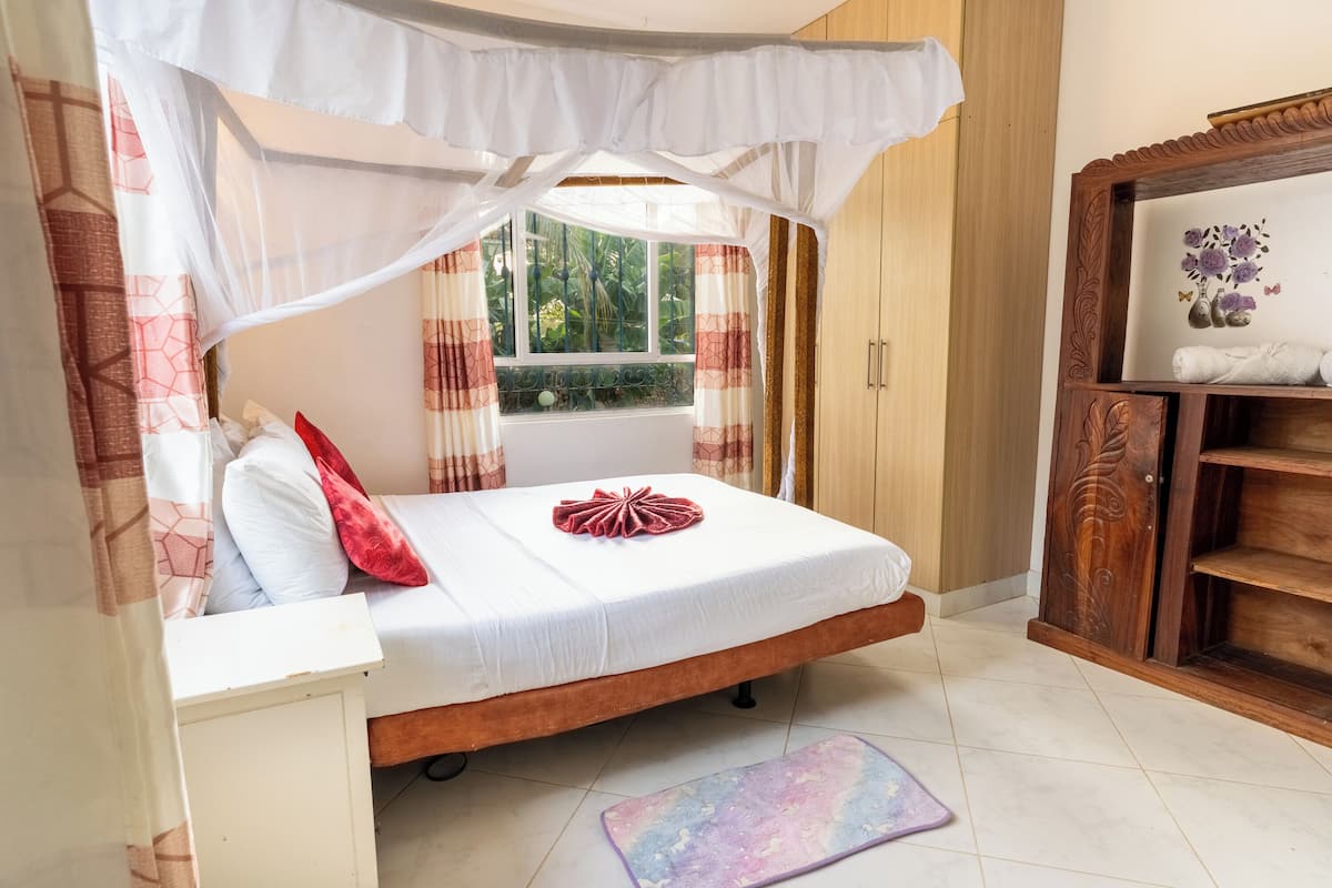 Un dormitorio con cama, mesita de noche, ventana con cortinas, armario de madera y una pequeña estantería con objetos decorativos.