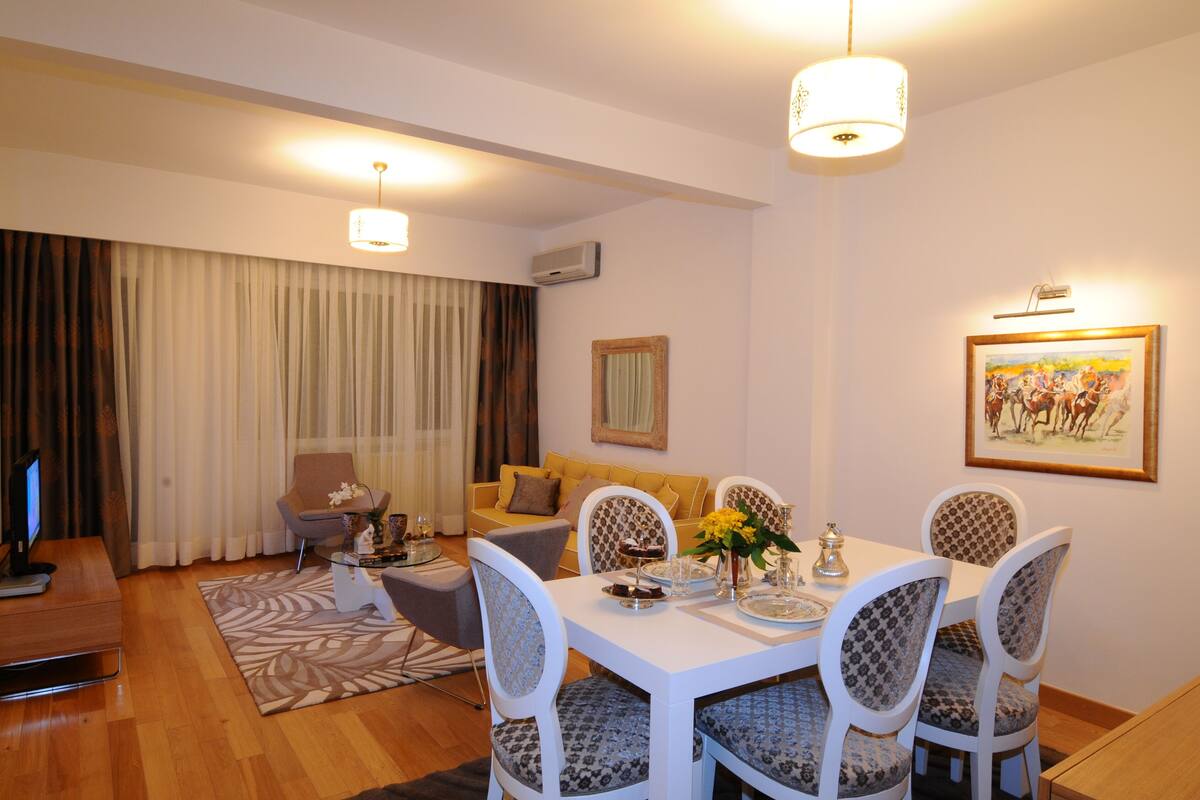 Un coin repas avec une table blanche, des chaises à motifs et un lustre. On aperçoit un canapé et un téléviseur à l’arrière-plan.