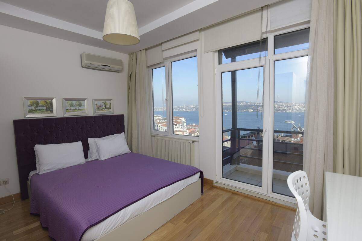 Une chambre avec un grand lit, une vue sur la ville depuis un balcon et un ventilateur de plafond.