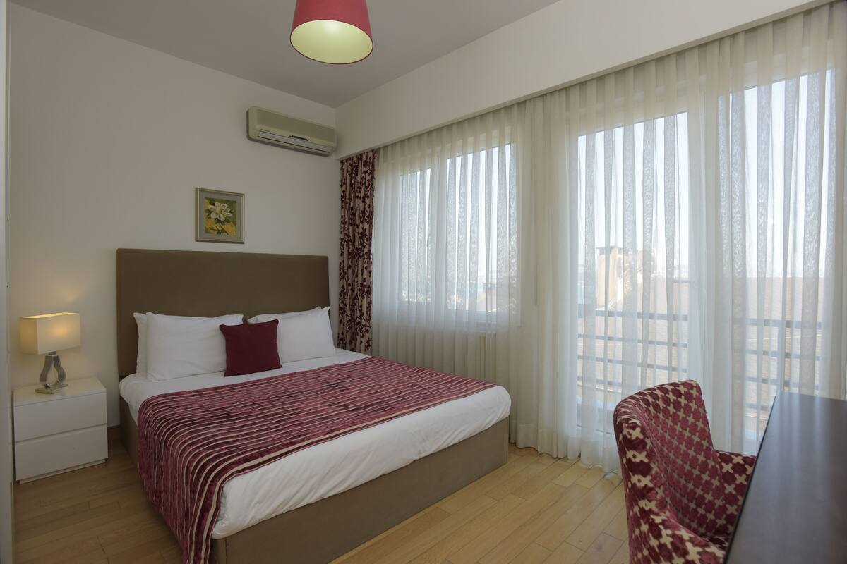 Une chambre d’hôtel avec un grand lit, une table de chevet avec une lampe, une chaise et une fenêtre avec des rideaux légers.