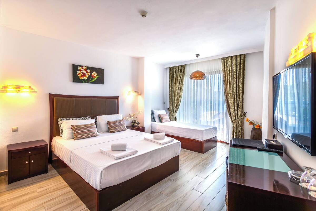 Hotellihuone, jossa on kaksi sänkyä, työpöytä ja tasatelevisio sekä suuri ikkuna, jossa on ohuet verhot.