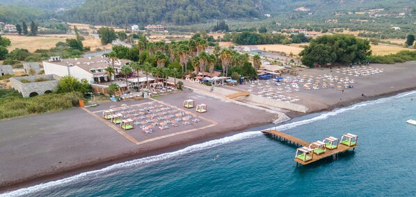 Vue aérienne d’un complexe hôtelier en bord de mer, avec un parking, des parasols de plage et une jetée en bois équipée de chaises longues.