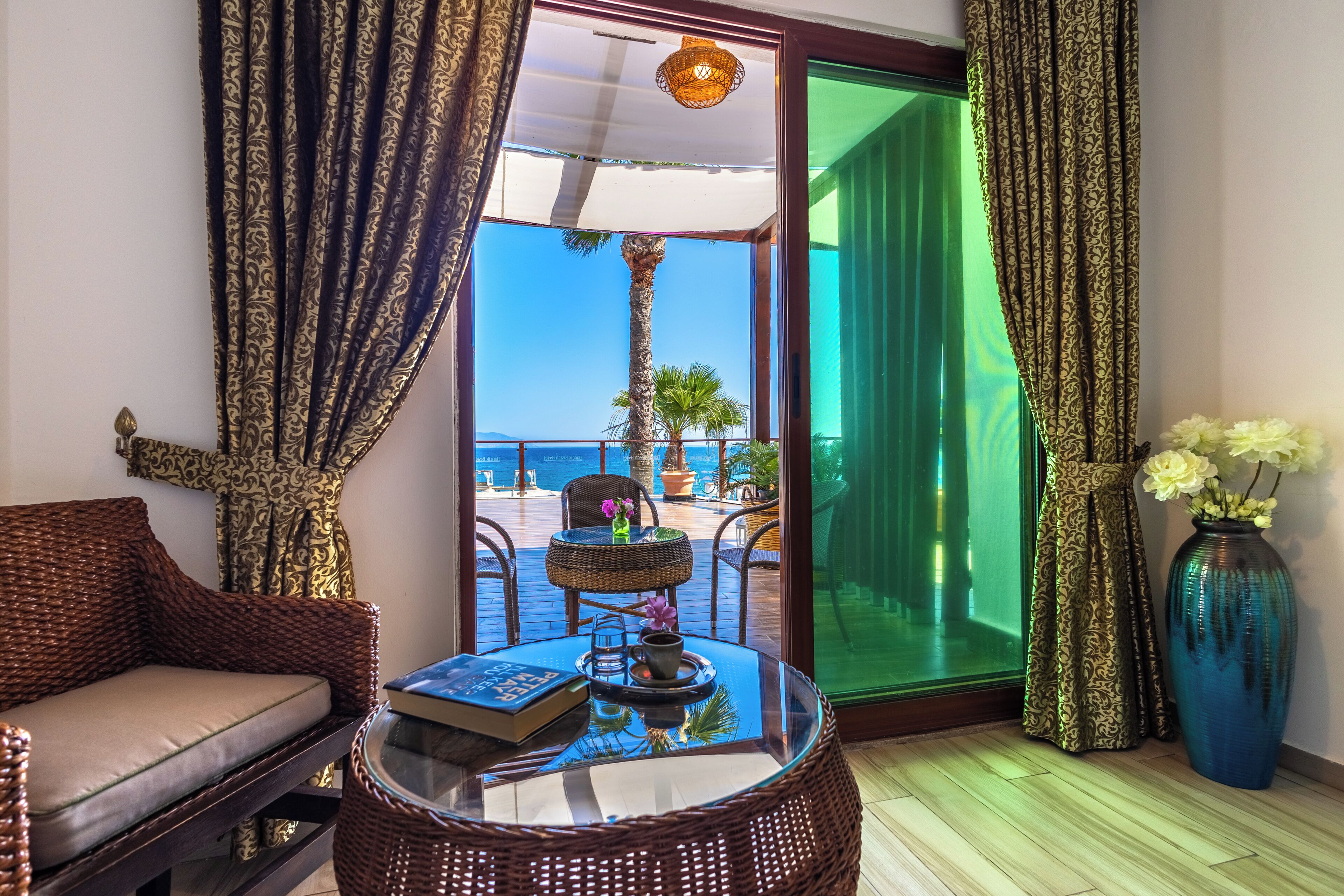 Una habitación con una silla de mimbre, una mesa redonda con un libro y una bandeja, un florero con flores y vista al mar a través de un ventanal.