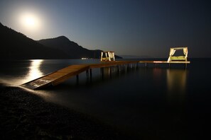 Une jetée en bois s’étendant sur une étendue d’eau calme, avec un lit placé sous un auvent, le tout entouré de montagnes et illuminé par une lune brillante.