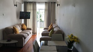 A living room with a sofa, a coffee table, a dining table with chairs, a flat-screen TV mounted on the wall, and a window with curtains.